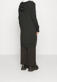 Dlouhý tmavě šedý pletený cardigan s kapucí, bočními rozparky a žebrovanými manžetami, nošený přes volné tmavě hnědé kalhoty a masivní černé boty.