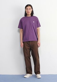 Paarse katoenen T-shirt met een klein wit logo op de borst, gecombineerd met bruine loszittende broeken en witte sneakers. Effen achtergrond.
