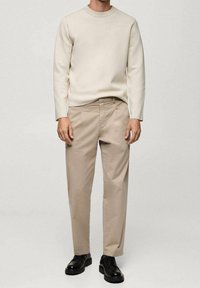 Homme portant un pull beige et un pantalon beige avec des chaussures en cuir noir, debout devant un fond clair uni.