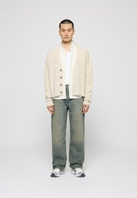 Beige strikket cardigan med kabelstrik, træknapper og ribbede manchetter, parret med lyseblå jeans og hvide sneakers.