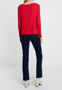 Rotes Strickoberteil mit langen Ärmeln und weitem Ausschnitt, kombiniert mit dunkelblauen geraden Jeans und cremefarbenen Ankle Boots. Einfaches Design, glatte Textur.