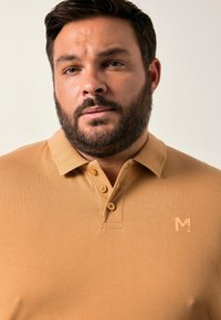 Vaaleanruskea polo-paita, jossa on kaulus, kolme nappia ja huomaamaton brändäys rinnassa. Kankaassa on teksturoitu, kudottu kuvio.