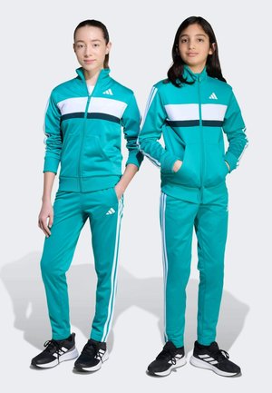 Twee meisjes in bijpassende turquoise trainingspakken van soepel materiaal. Elk heeft witte strepen en een Adidas-logo op de borst en broek. Zwarte sneakers.