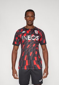 Červený a čierny futbalový dres s geometrickým vzorom, okrúhlym výstrihom, krátkymi rukávmi a logom "INEOS" na prednej strane. Čierne šortky.