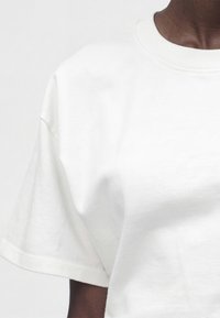 Κοντινό πλάνο ενός ατόμου που φοράει απλό λευκό κοντομάνικο T-shirt με στρογγυλή λαιμόκοψη σε φωτεινό γκρι φόντο.