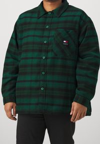 Camicia in flanella a quadri verde e nera, con colletto, chiusura a bottoni e taschino sul petto con patch logo. Texture morbida, vestibilità rilassata.
