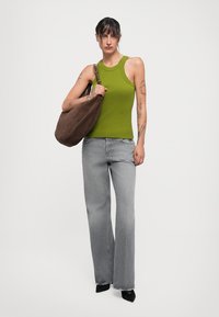 Kvinna med tatueringar som bär en grön ärmlös topp och vida grå jeans, håller en stor brun axelväska.