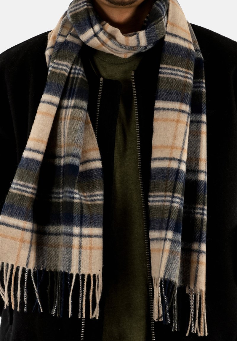 Gros plan d'une personne portant une veste noire, une chemise olive, et une écharpe à carreaux beige, bleue et vert foncé avec des franges.