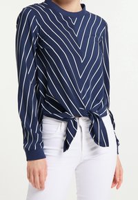 Marinebluse mit weißen vertikalen Streifen, runder Halsauschnitt, langen Ärmeln und einem gebundenen Detail vorne. Hergestellt aus leichtem Material.
