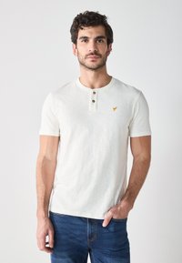 Camisa de manga corta color beige claro con cuello redondo, cierre de tres botones y un pequeño logotipo bordado en el lado izquierdo del pecho. Textura lisa.