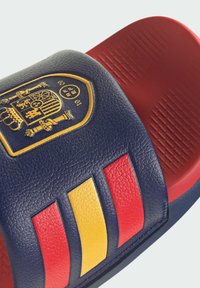 Sandalia tipo chancla en azul marino y rojo con suela texturizada, que presenta el escudo de armas de España y franjas rojo-amarillo-rojo en la correa.