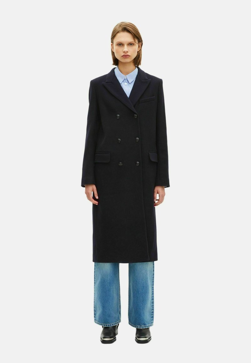 Dubbelzijdige marine-wollen jas met gekante kraag en zijzakken, gedragen over een lichtblauw shirt en wijde jeans, afgewerkt met zwarte schoenen.