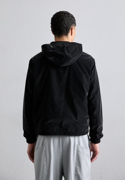 Casaco leve preto com capucho, apresentando punhos elásticos e um corte direito. O tecido tem uma textura suave e mate. Usado sobre calças cinza claro.