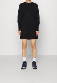 Svart stickad klänning med rund halsringning, puffärmar och texturerade manschetter. Tillsammans med mörka sportskor och vita ankelsockor.