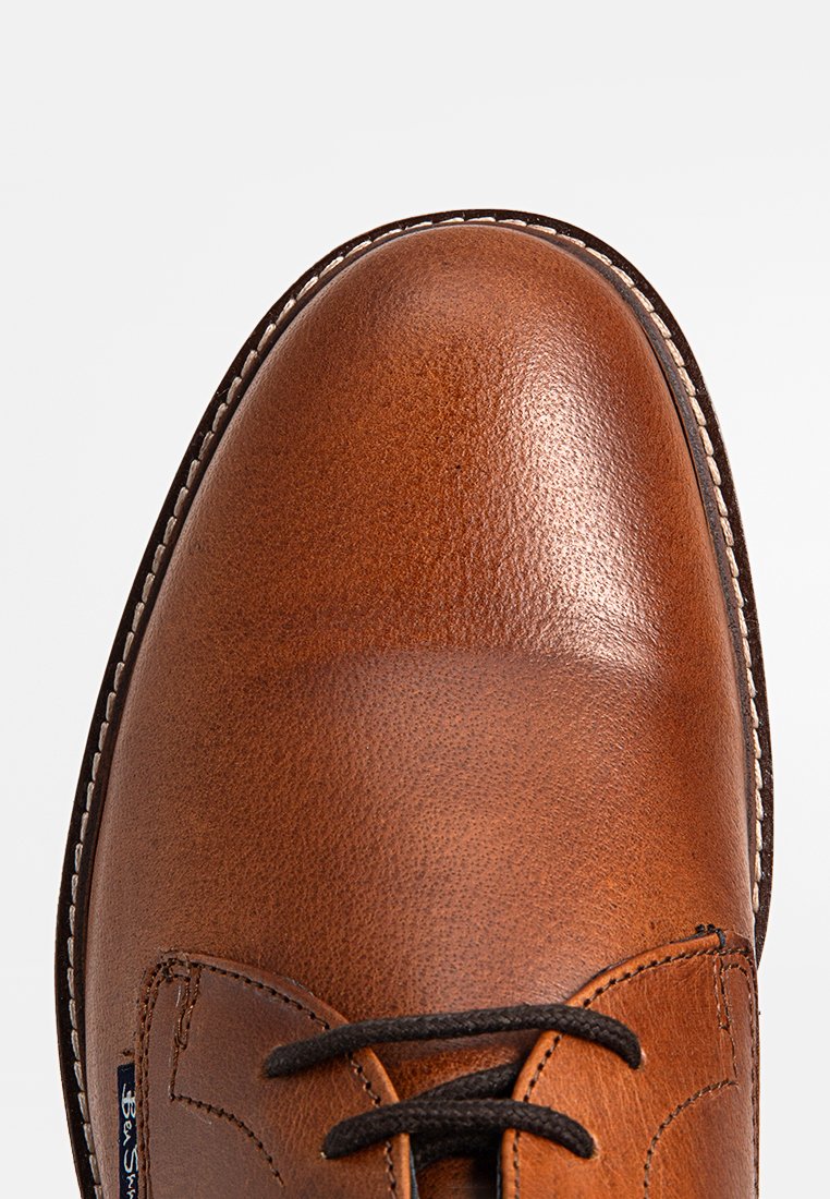 Zbliżenie na wypolerowany brązowy skórzany nos buta z detalicznym przeszyciem i czarnymi sznurowadłami na neutralnym tle.