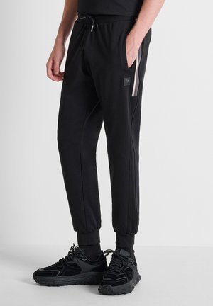 Uomo che indossa pantaloni jogger neri con polsini elastici e banda laterale, abbinati a sneakers nere chunky su sfondo semplice.