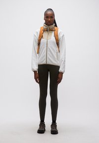 Γυναίκα που φοράει μπεζ και λευκό μπουφάν με φερμουάρ, ελαφρώς πράσινο κολάν, παπούτσια πεζοπορίας και πορτοκαλί σακίδιο πλάτης, στέκεται μπροστά σε μονόχρωμο φόντο.