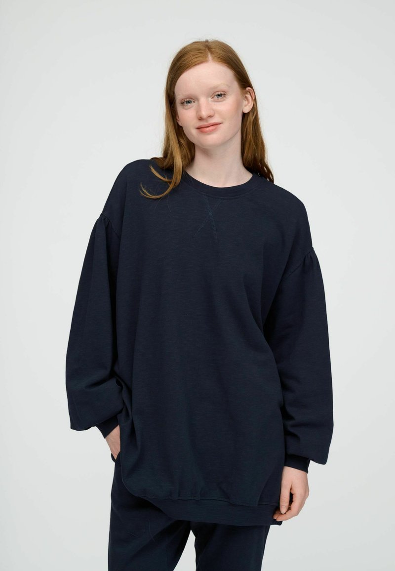 Jeune femme aux cheveux roux portant un sweat-shirt ample bleu marine foncé et un pantalon, debout devant un fond clair uni.