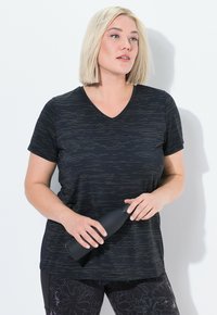 Blondýnka v černém vzorovaném sportovním tričku a legínách, která drží černou lahev s vodou, dívá se do strany na bílém pozadí.