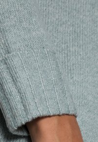 Pull tricoté bleu clair avec poignets côtelés, montrant un matériau texturé et doux ainsi que des variations subtiles de couleur sur toute la surface.