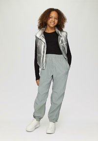 Zilveren opblaasvest over een zwarte gestructureerde trui, gecombineerd met lichtgrijze broek met elastische taille en witte sneakers. Krullend haar.