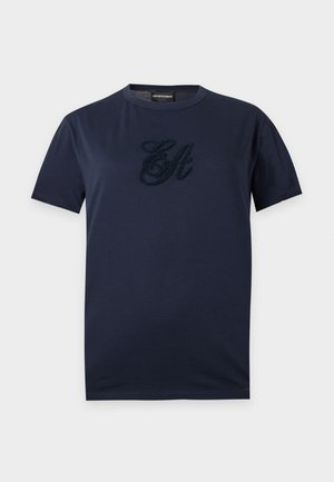Femme portant une chemise bleu marine à manches courtes et un pantalon beige à jambes larges, debout les mains dans les poches sur un fond uni.