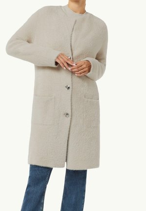 Femme portant un long manteau beige duveteux boutonné par-dessus un jean bleu, ajustant le bouton supérieur avec les deux mains.
