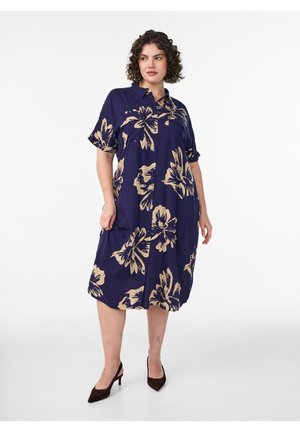 Naiste, kes kannab tumesinist põlvikuni ulatuvat särgikleit suurte beežide lillemustritega ja musti kõrge kontsaga kingi, seisab valge tausta ees.