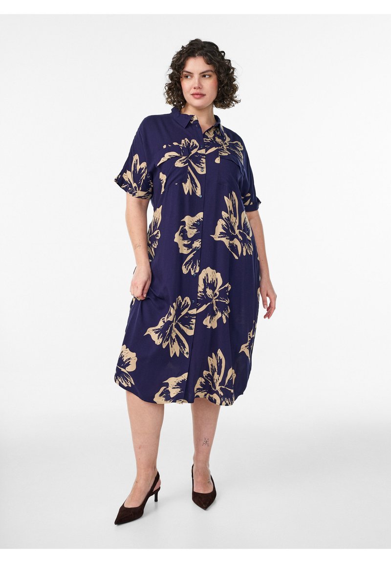 Frau trägt ein knieumspieltes, marineblaues Hemdblusenkleid mit großen beigen Blumenmustern und schwarzen High Heels, steht vor einem weißen Hintergrund.