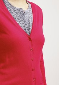 Fuksia cardigan, jossa on V-pääntie ja viisi nappia, kerrostettu kuvioidun tummansinisen ja valkoisen paidan päälle. Sileä pinta ja istuva muotoilu.