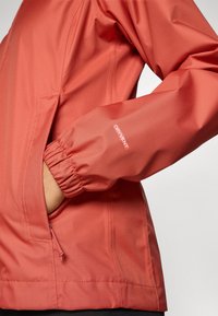 Water-resistant orange jacket with elastic cuffs, side pocket, and logo detail. Smooth texture and casual design for outdoor wear.