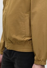 Καφέ μπουφάν Barbour με ελαστικές μανσέτες και τελείωμα, φορεμένο με μαύρο παντελόνι, που δείχνει λεπτομέρεια πλαϊνής τσέπης με κεντημένο λογότυπο Barbour.