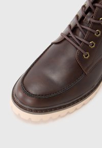 Bota de cuero marrón con punta redonda, ojales de metal y cordones marrones. Presenta una suela de goma blanca texturizada con bordes serrados.