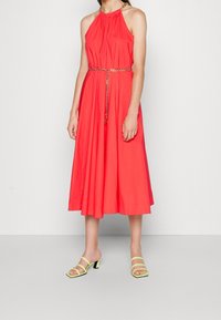 Femme portant une robe midi sans manches corail avec une ceinture en chaîne et des sandales à talons à lanières vert citron, se tenant sur un sol gris.