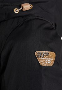 Černá tkanina s texturovaným povrchem, tvořená hnědou koženou nášivkou s vyraženým logem a stahovací sponou nahoře.