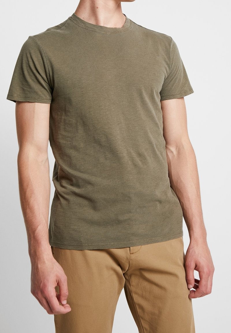 Homme portant un t-shirt à manches courtes vert olive et un pantalon marron clair, debout devant un fond clair uni.