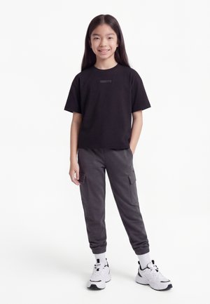 Usmievajúce sa dieťa s dlhými tmavými vlasmi oblečené v čiernom tričku, tmavých cargo teplákoch, bielych ponožkách a bielych teniskách, stojace s jednou rukou v vrecku.