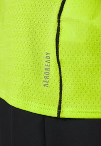 Fluorescerande gul atletisk tröja gjord av texturerat tyg, med en svart söm och "AEROREADY"-logotyp i grått på den nedre vänstra sidan.