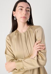 Blusa di raso beige con scollatura arricciata, maniche lunghe e polsi con bottoni. Modella con orecchini rotondi in tonalità argento. Texture liscia.
