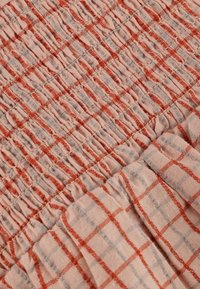 Světlo, jemná látka s červeným a šedým kostkovaným vzorem, s nařasenou texturou. Rukáv s volánem přidává dimenzionální detail.