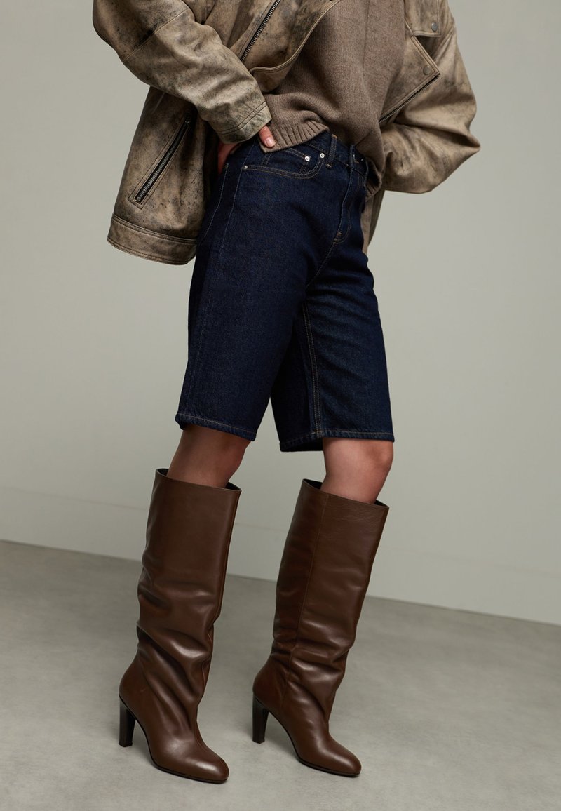 Personne portant un short en jean noir, des bottes hautes à talons bruns, un pull brun et une veste en cuir brun, les mains sur les hanches.