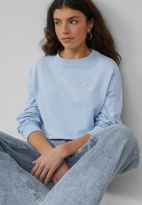 Jasnoniebieska bluza z miękkiego materiału z okrągłym dekoltem, ściągaczami przy rękawach oraz małym haftowanym logo na piersi. Połączona z jasnymi dżinsami.