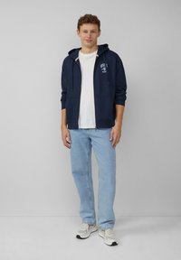 Felpa navy con zip, cappuccio con cordini, tasche frontali e logo bianco. Abbinata a jeans di denim azzurro chiaro e scarpe da ginnastica chiare.