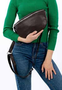 Ruda odinė per petį nešama rankinė su lenkta forma, sidabriniu užtrauktuku ir reguliuojamu dirželiu, laikoma asmens, dėvinčio žalią ribotą megztinį.