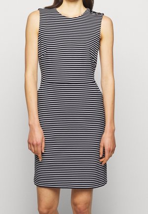 Vrouw draagt een mouwloze, knie-lange jurk met zwarte en witte horizontale strepen en een knoopdetail op de linker schouder.
