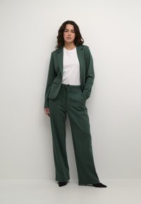 Groene op maat gemaakte blazer met bijpassende losse broek. Zacht materiaal, slanke revers op de jas, broek met een koordsluiting in de taille, en zwarte puntige schoenen.