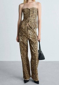 Žena v topu bez ramínek s leopardím potiskem a odpovídajících kalhotách, držící černou kabelku, stojící na šedé podlaze proti světlé zdi.