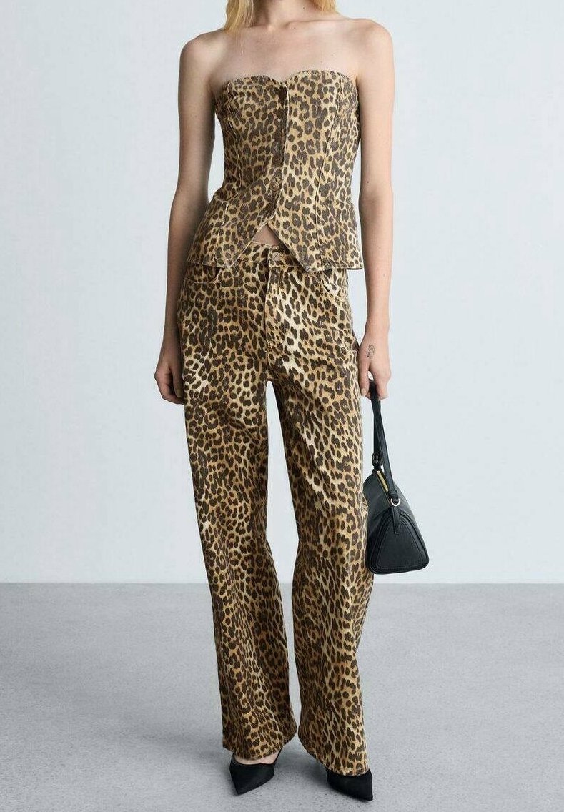 Žena v topu bez ramínek s leopardím potiskem a odpovídajících kalhotách, držící černou kabelku, stojící na šedé podlaze proti světlé zdi.