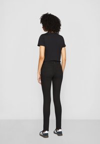 Mujer con cabello rizado que lleva una camiseta negra, jeans ajustados negros y zapatillas blanco y negro, de pie frente a un fondo gris liso.