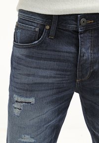 Close-up of dark blue denim jeans with distressed details on thigh and visible button and belt loops, worn with white knit sweater.